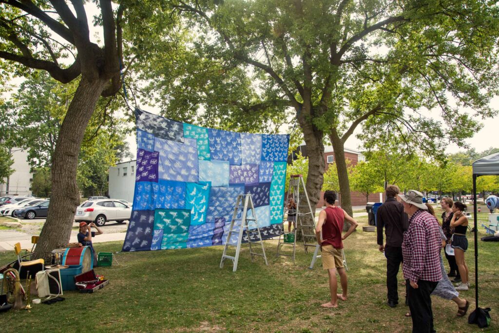 In a park between two giant trees a big quilted blanket with screen printed birds is hoisted up. People look at it with excitement. 